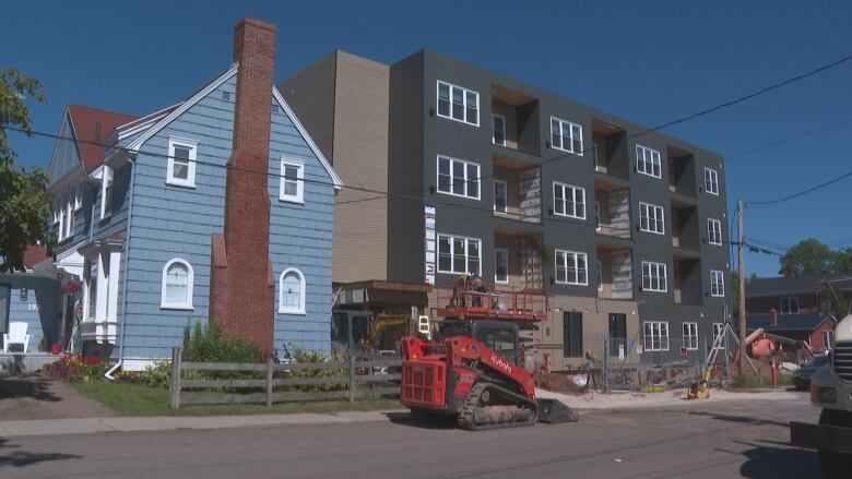 An apartment building with construction equipment and fencing around it stands next to a blue house. 