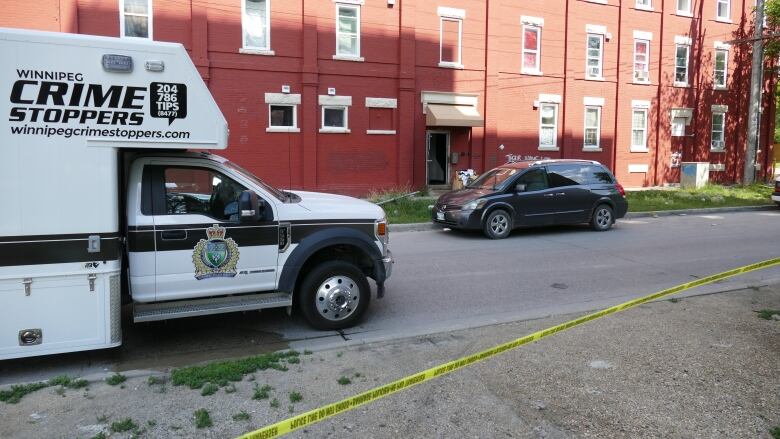 Yellow police tape is in the foreground with a black-and-white police van that says Winnipeg Crime Stoppers. A red brick apartment building is in the background.