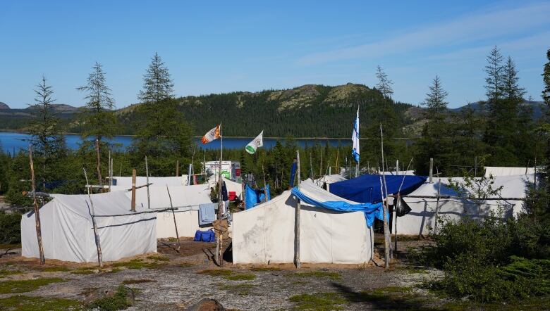 A group of Innu tents, some flying Innu flags, are grouped against a scenic backdrop of water and mountains.