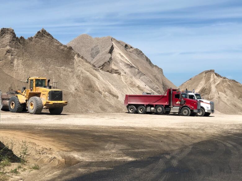 A tractor is seen with two dump trucks against a backdrop of a giant pile of crushed stone.