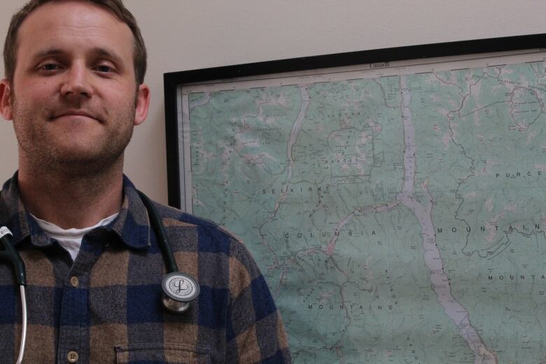 A doctor smiles in front of a map. He is wearing a checked shirt and has a stethoscope on.