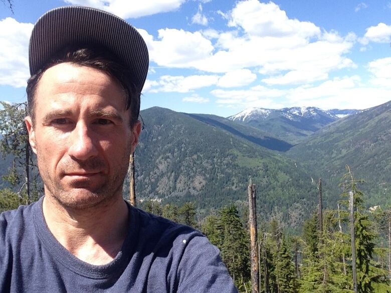 A man wears an upturned baseball hat and a blue T-shirt as he poses in front of mountains.