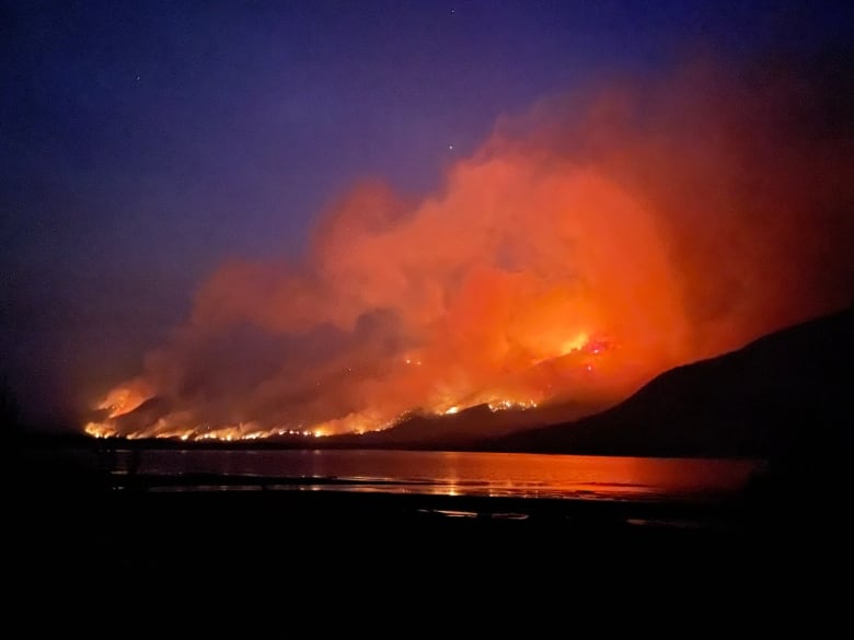 It's nighttime. Flames glow on a mountain side with massive clouds of smoke floating upward. The surface of a lake is in the foreground; the colour of the flames is reflected on the water.