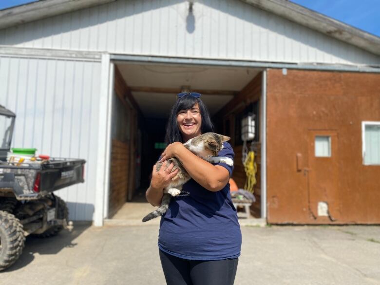 A woman stands in front of a barn smiling. She holds a cat in her arms.