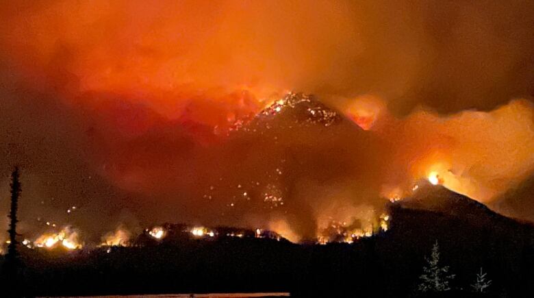 It's nighttime. Flames of orange spot a mountain, and smoke clouds haze the area.