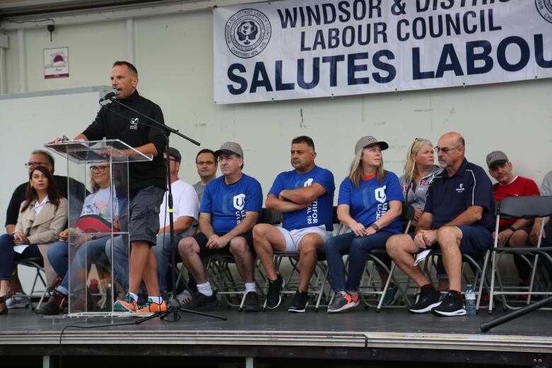 A man wearing a black jacket delivering a speech with people sitting in chairs behind him