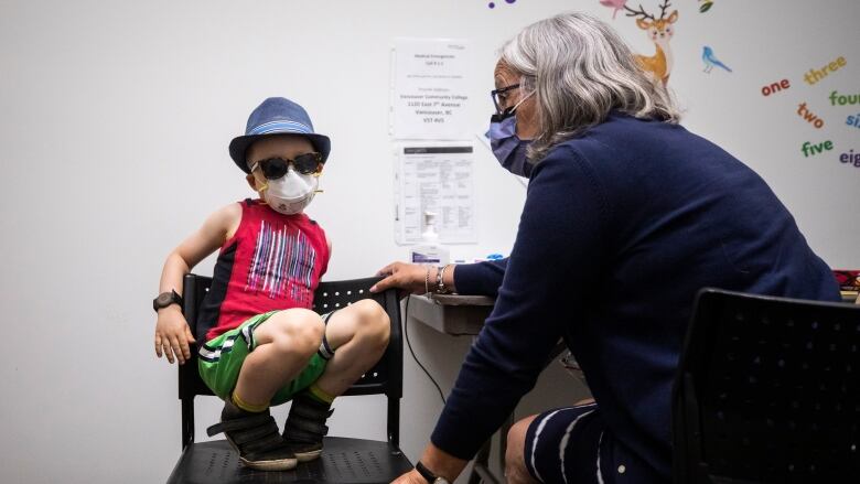 A young boy crouches on a chair in a vaccination clinic in Vancouver, wearing sunglasses, an N95 mask and a fedora.