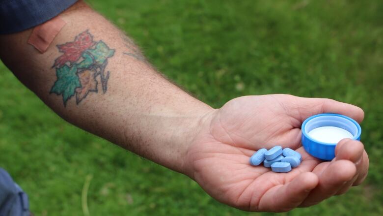 A man's tattooed arm is extended in front of green grass. Blue pills rest on the hand.