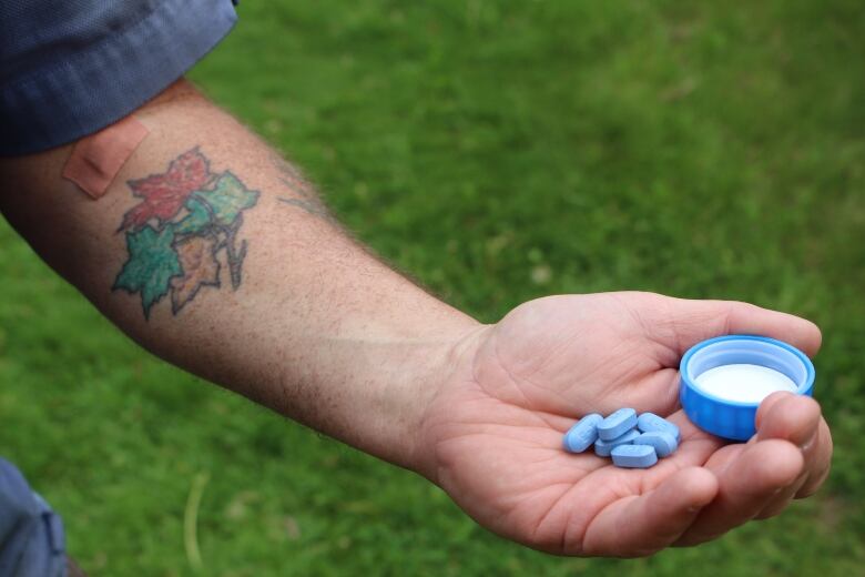 A man's tattooed arm is extended in front of green grass. Blue pills rest on the hand.