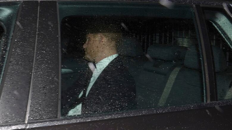 A man with red hair, wearing a black suit jacket and white suit, seen through a rain-covered window in the back seat of black car. 