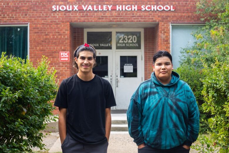 Two young men stand in front of a school building with a sign that says Sioux Valley High School.