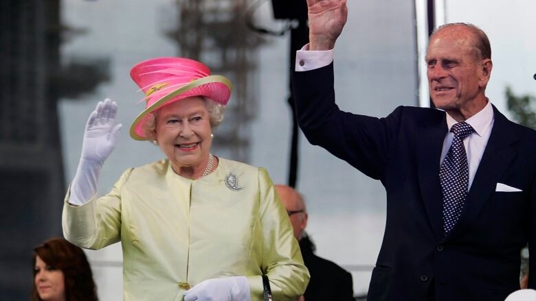 A woman in a pale yellow dress and a pink hat and a man in a dark suit stand on a stage, waving.