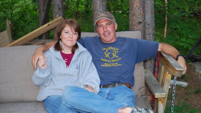 A woman in a grey sweater sits beside a man with a blue shirt on a bench outside.