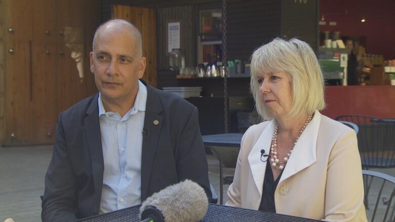 Pete Fry and Adriane Carr look pensive as they sit at a table. Fry is a Black man wearing a blue shirt and blazer. Carr is a white woman with short white hair.