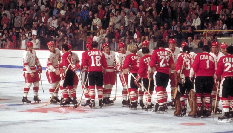 Two lines of players shake hands on the ice. The Canadians, closer to the camera, wear red sweaters with white trim, while the Soviets wear white sweaters with red trim.