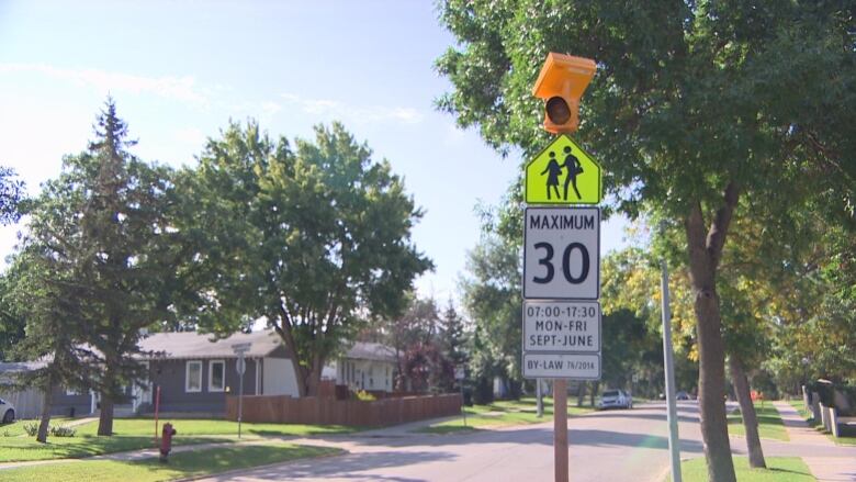 An amber warning light sits atop a post on a treed street, above signs indicating a school zone and a 30 kilometre per hour speed limit from 7 a.m. to 5:30 p.m., Monday to Friday, September to June.