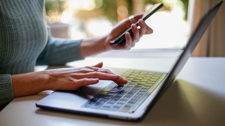 Hands type at a laptop and hold a cellphone