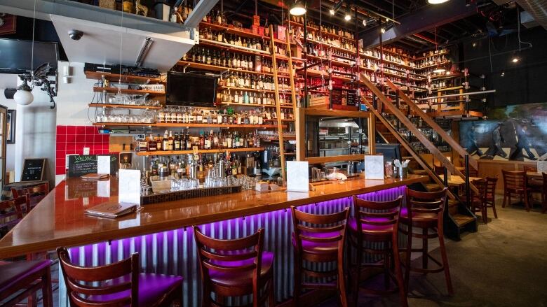 A bar with purple neon lighting underneath it, and dozens of bottles behind it. A ladder and stairs lead up to a level with more bottles.