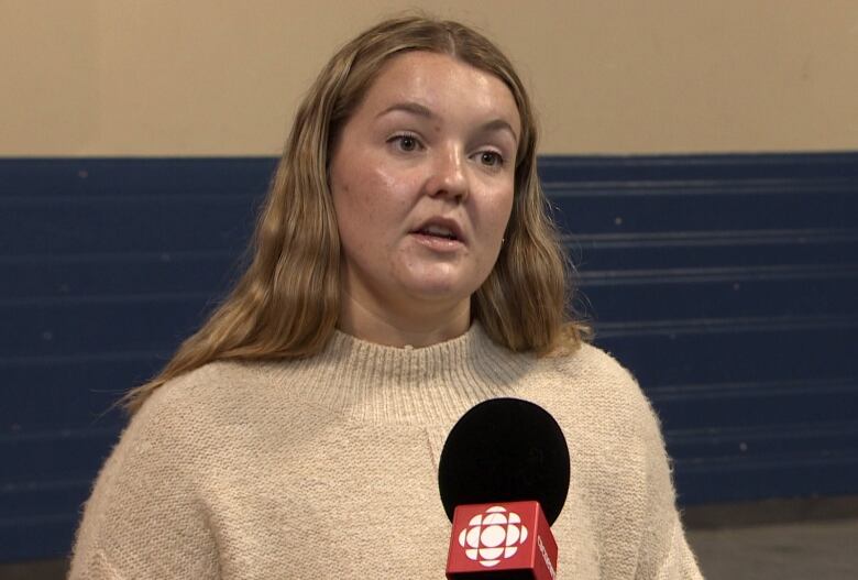 A chest-up shot of a person in a beige sweater with long blonde hair speaking into a microphone.