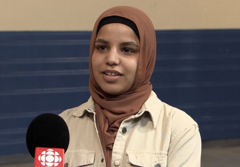 A person wearing a white jacket and a brown hijab speaks into a microphone.