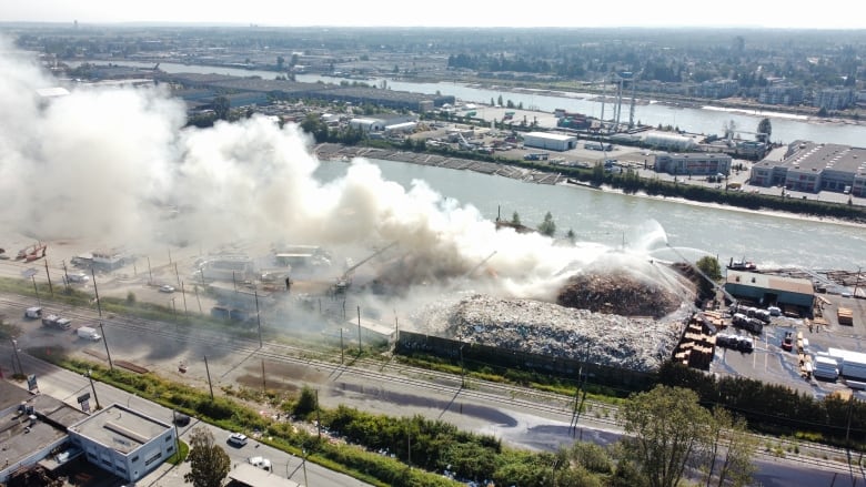 An aerial shot shows a plume of smoke rising from a landfill, surrounded by industrial buildings. Large fire engine attachments are seen attempting to douse the fire.