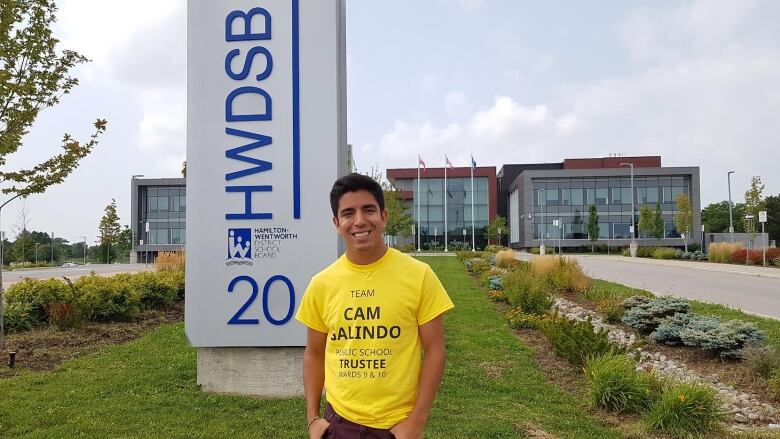 A man in a yellow campaign shirt stands in front of a building. 