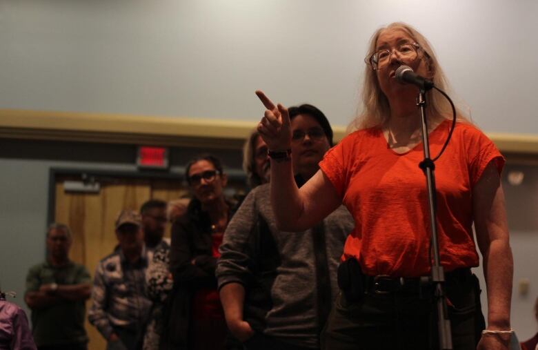 A woman stands at the mic pointing a finger. Behind her is a long line of people waiting.