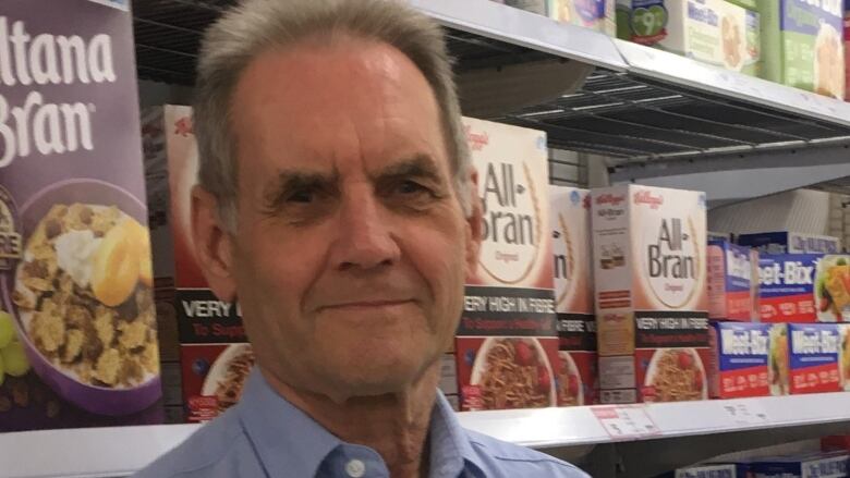 A short-haired individual standing in front of a shelf of cereal boxes, with a serious expression on his face. 