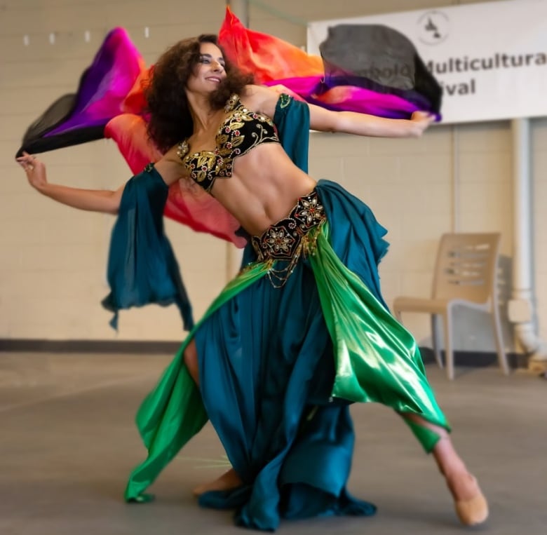 A belly dancer performs a dance routine. She has a smile on her face, and has a series of cloths flowing behind her head coming from either hand.