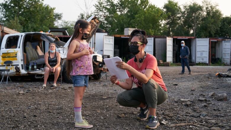 A little girl stands with a mask on. A woman crouches down next to her, holding sheets of paper. 