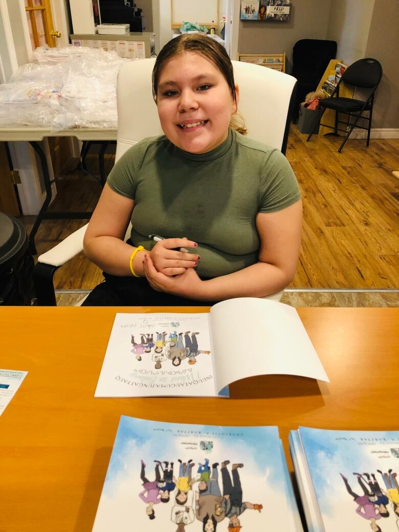 A woman holding a pen smiles at the camera. In front of her, a book is open to its first page.