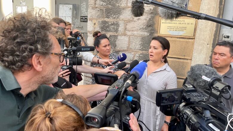 A woman speaks into a row of microphone as reporters listen.