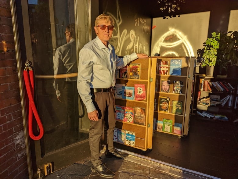 A man in sunglasses and great shoes stands next to a set of bookshelves.