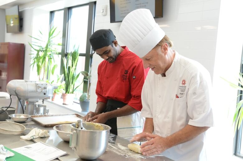 A chef in uniform stands with a student rolling dough in a commercial kitchen 