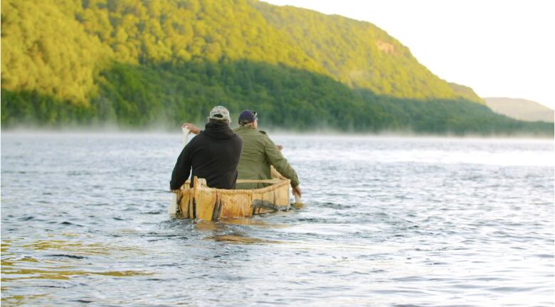 Two men on the water  in a birchbark canoe with a hill of green in front of them.