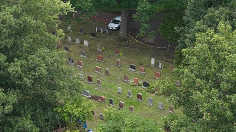 Evidence markers and crime tape are seen near a silver car and tombstones.