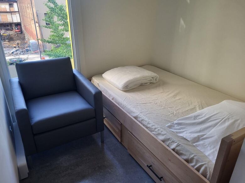 A black chair and single bed with white sheets and a pillow by a window in the new shelter.