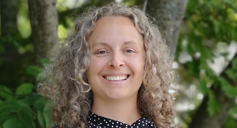 A woman with grey curly hair is standing outside in a black dress with small white dots.