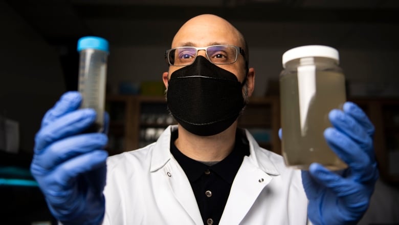 A man in a facemask holds up a jar and a test tube with murky liquids inside them.