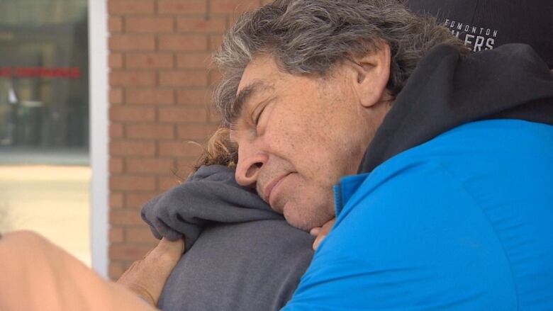 a man in a blue jacket hugs another person with a brick wall in the background