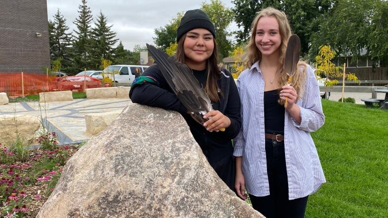 Two smiling young women stand together, each holding a feather.