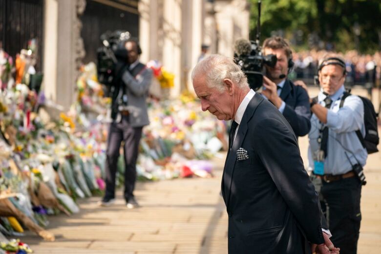 A grey haired man stand solemnly before flowers laid outside Buckingham Palace , while camera people stand behind him taking photos. 