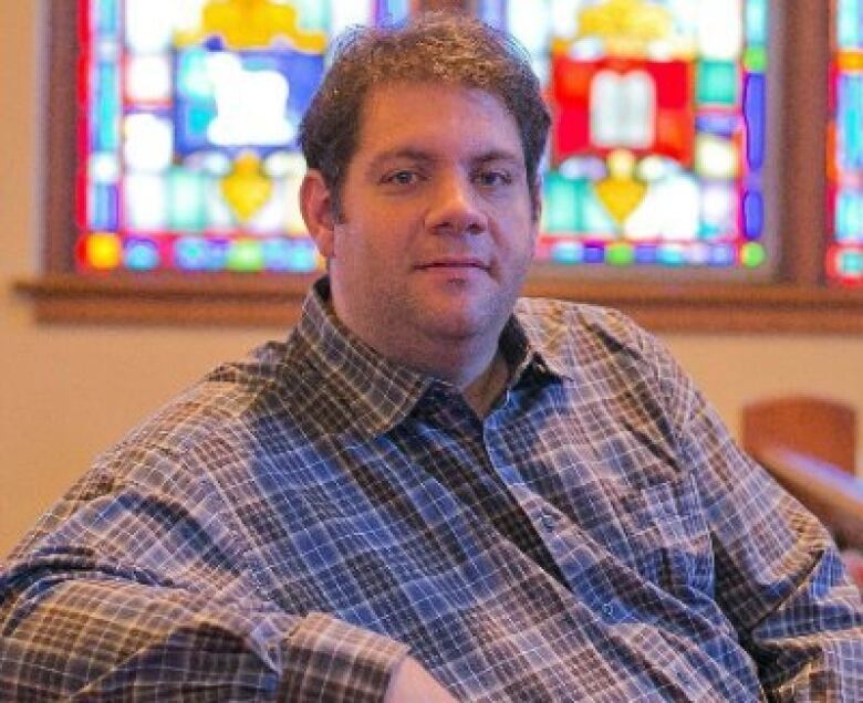 A sitting man wearing a checkered shirt faces the camera, with stained glass windows in the rear 