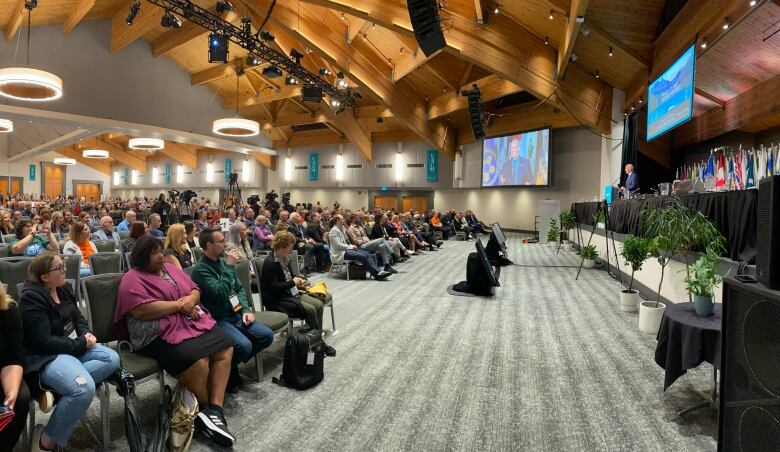 Dozens of people are seen at a conference listening to a white man speak.