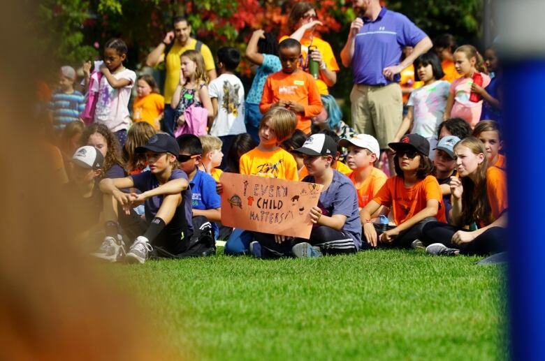 Dozens of kids wearing orange shirts and holding signs sit in a grassy field.