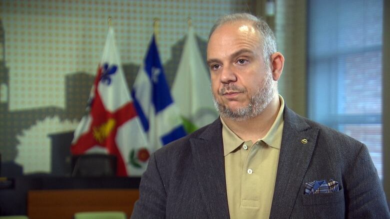 A man wearing a polo shirt and jacket stands in front of the Montreal and Quebec flags. 