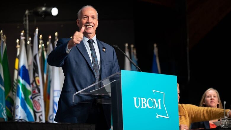 John Horgan, an old white man with a white moustache, gives a thumbs up as he stands at a podium.