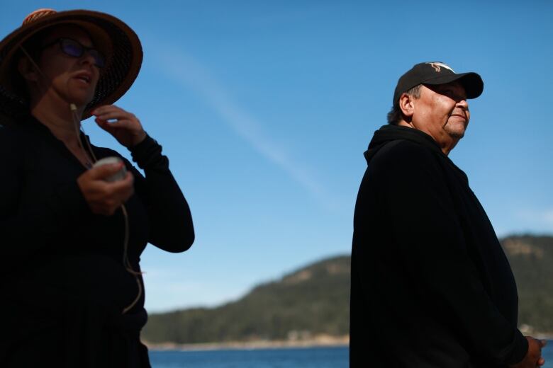 Two Indigenous people gesture, as they stand in the backdrop of an island.