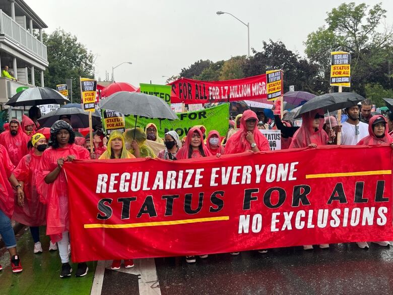 Many people wearing raincoats and holding umbrellas stand behind a red banner.