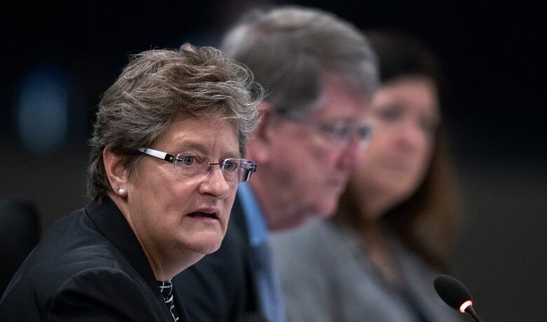 A panel of people can be seen speaking in front of microphones. Two are blurry, while the woman in the foreground has short gret hair and glasses.